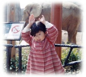 （写真）動物園のゾウの前で、頭の上に手を乗せ、ゾウのポーズをとる幼い夏衣